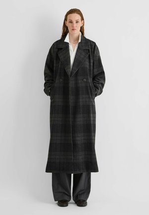 Un mannequin se tient devant un fond uni, vêtu d’un long manteau sombre à carreaux et à double boutonnage, par-dessus une chemise blanche et un pantalon sombre.