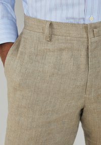 Uomo con pantaloni di lino beige e una camicia blu a righe bianche, con una mano parzialmente in tasca, ritratto fino alla vita e a metà coscia.