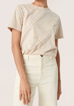 Persona vistiendo una camiseta de manga corta beige con patrones de líneas geométricas, metida dentro de unos pantalones de talle alto color blanco roto, de pie contra un fondo liso.