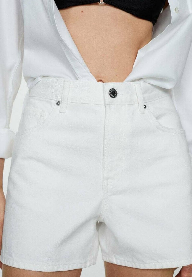 Shorts de mezclilla blanca con tiro alto, que cuentan con un cierre de botón frontal, trabillas para cinturón y bolsillos laterales, exhibidos con una camisa blanca desabotonada.