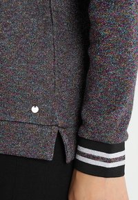 Jersey multicolor con una textura brillante, que presenta puños negros con rayas blancas y un pequeño acento de logo plateado.