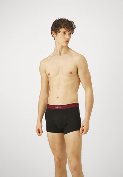 Boxers noirs avec une ceinture bordeaux arborant un texte logo blanc. Le tissu semble lisse et ajusté. Le modèle est torse nu.