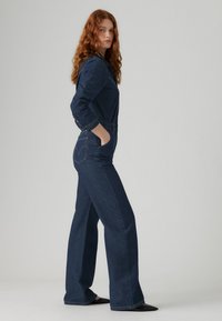 Combi-short en denim bleu foncé avec manches longues, une fermeture à boutons sur le devant, des poches latérales et un design à jambe large. Caractérisé par une couture signature sur la poche arrière.