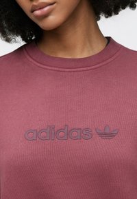 Μπορντό φούτερ με ριμπ λαιμόκοψη και υφασμένο λογότυπο "adidas" και έμβλημα trefoil σε πιο σκούρο τόνο, με λείο φινίρισμα υφάσματος.