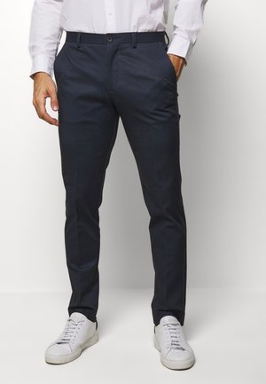 Homme portant un pantalon slim à carreaux bleu foncé, une chemise blanche à manches longues et des baskets blanches, debout les mains partiellement dans les poches.