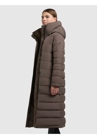 Palton lung căptușit, de culoare maro, cu guler înalt, glugă și închidere cu capse/butoane. Prezintă un croi structurat cu model matlasat orizontal.