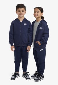 Haut à zip bleu marine et pantalon de survêtement assorti en tissu doux. Chaque pièce arbore le logo Nike en blanc. Les baskets sont noires et grises.