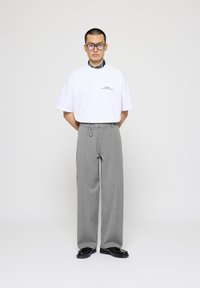 Camiseta blanca de gran tamaño con texto bordado, combinada con pantalones grises de cintura alta que tienen un lazo para cinturón y un detalle de llavero. Zapatos negros.
