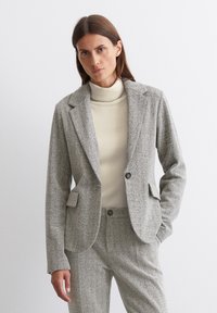 Grauer Hahnentrittmuster-Blazer mit Reverskragen und Einzelknopfverschluss. Kombiniert mit einem beigefarbenen gerippten Rollkragenpullover. Strukturiertes Material, taillierte Passform.