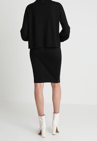 Svart kort jacka över en åtsittande svart kjol, kombinerat med vita anklarstövlar. Slätt tyg, enkel design och rena linjer.