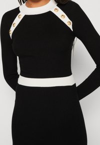 Robe pull noire à côtes avec encolure et taille à côtes blanches. Présente des boutons dorés sur les épaules. Design ajusté.