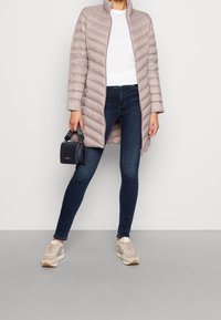 Cappotto imbottito di un beige chiaro con cuciture verticali, colletto alto, abbinato a jeans in denim scuro e sneaker beige, mentre tiene una piccola borsa nera.