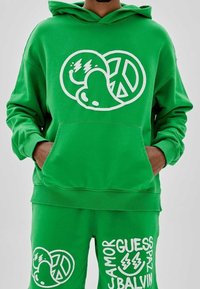 Persona vestida con un conjunto de sudadera verde y pantalones cortos, decorado con símbolos de paz, corazón y carita sonriente en blanco, además de texto en negrita que incluye "AMOR", "GUESS" y "J BALVIN".
