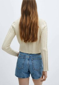 Cremfarbener Strickpullover mit einem cropped Design, kombiniert mit hoch geschnittenen blauen Denim-Shorts mit Gesäßtaschen.