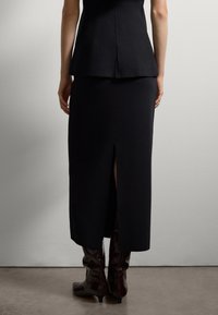 Schwarzes, tailliertes Oberteil mit einem Peplum-Saum, kombiniert mit einem langen schwarzen Rock mit einem Rückenschlitz und knielangen, glänzenden bordeauxroten Stiefeln.