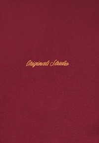 Bordo spalvos audinys su geltonai išsiuvinėtu tekstu „Originals Studio“. Lygus paviršius su švelniu tamprumu.