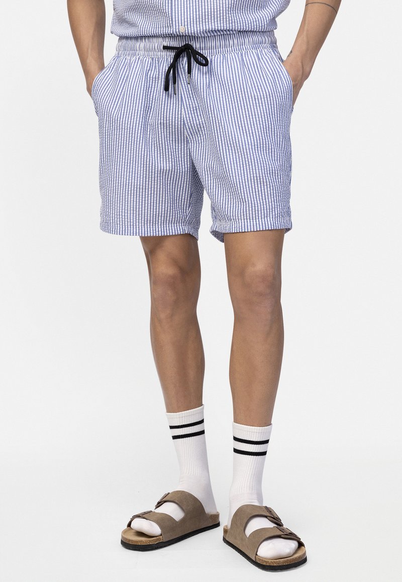 Άνδρας που φοράει μπλε και λευκές ριγέ βερμούδες με μαύρο κορδόνι, άσπρες κάλτσες με μαύρες ρίγες και καφέ σανδάλια με αγκράφα, όρθιος.