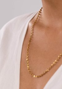 Goldene Kettenhalskette mit runden Gliedern, die auf nackter Haut liegt, vor einem weißen Stoffhintergrund. Glatte Textur und polierte Oberfläche.