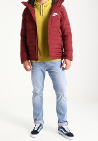 Burgundy quiltad jacka med huva, grå foder och Nike-logotyp; bärs över en senapsgul hoodie, i kombination med ljusblå jeans och marinblå Vans-skor.