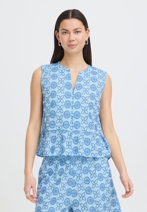 Kvinde med langt mørkt hår iført ærmeløs lyseblå top og matchende shorts med mørkeblå blomsterprint, stående mod en hvid baggrund.