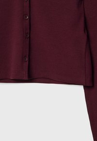 Gros plan d'un cardigan en maille côtelée bordeaux avec des boutons devant et des manches longues sur un fond uni.