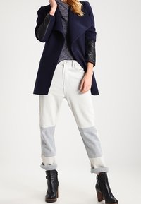 Femme portant un manteau bleu marine avec des manches en cuir noir, un pull en maille violet, un pantalon blanc avec des renforts gris aux genoux, et des bottines à talons noires.