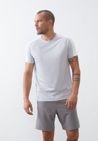 Helles graues Sport-T-Shirt mit kurzen Ärmeln und einem strukturierten Muster, kombiniert mit grauen Shorts. Zeigt sichtbare Tattoos am Arm.