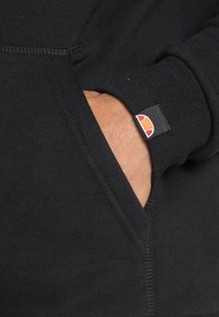 Sudadera negra con capucha que presenta un bolsillo frontal y puños acanalados. Un detalle notable incluye un pequeño parche con un logotipo en naranja y blanco.