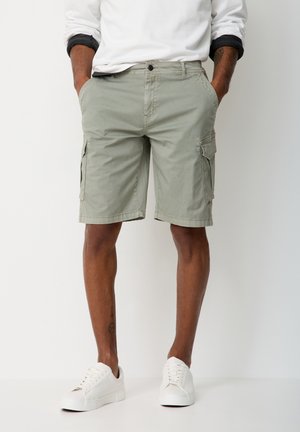 Mann trägt hellgrüne Cargo-Shorts, weiße Turnschuhe und ein weißes Langarmshirt, die Hände in den Taschen, steht vor einem einfarbigen Hintergrund.