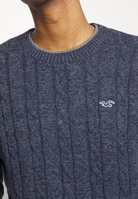 Pull en maille torsadée bleu marine avec un col rond, présentant une délicate bordure grise et un petit logo brodé sur la poitrine.