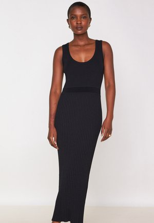 Femme portant une robe maxi noire sans manches à côtes, se tenant devant un fond clair, regardant devant elle avec une expression neutre.