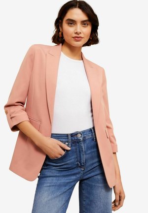 Donna con capelli corti e scuri, indossa orecchini a cerchio, un blazer rosa pallido con maniche arrotolate, una maglietta bianca e jeans blu, mentre posa con una mano in tasca.