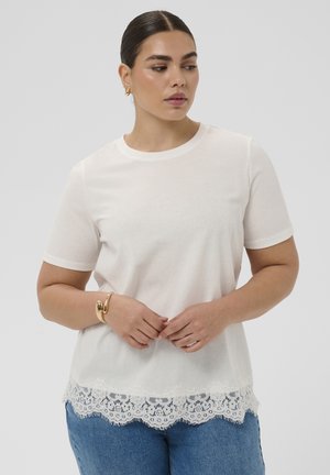 Frau mit dunklen Haaren, die ein weißes Kurzarmshirt mit Spitzenabschluss, blaue Jeans und ein goldenes Armband trägt, schaut nach links vor einem grauen Hintergrund.