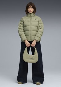 Kvinna i lätt grön vadderad jacka och vida mörka jeans, som håller en matchande lätt grön handväska, stående mot en grå bakgrund.
