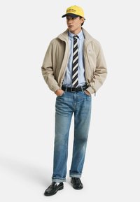 Giacca beige, camicia a righe blu, cravatta a righe, jeans azzurri chiari, scarpe nere, cappellino giallo e blu navy. Strati casual con diverse texture.