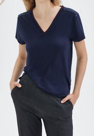 Vrouw draagt een marineblauw T-shirt met V-hals en korte mouwen, ingestopt in donkergrijze, gestructureerde broek met handen in de zakken.