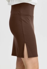 Jupe moulante brune en tissu doux, dotée d'une taille haute et d'une fente à l'arrière pour faciliter les mouvements. Design simple sans motifs supplémentaires.