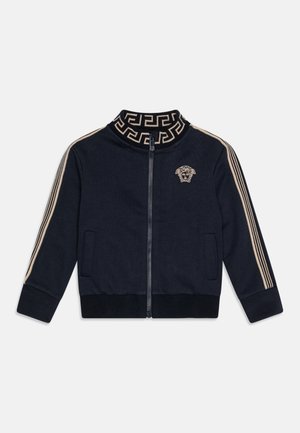 Marineblaue Jacke mit Reißverschluss, mit beigem griechischem Schlüsselmuster am Kragen, beigen Streifen an den Ärmeln und einem beigen Medusa-Logo auf der Brust.