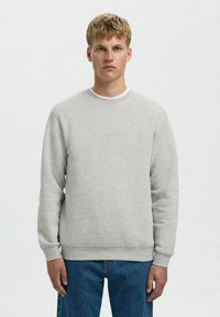 Sweatshirt gris clair avec col rond et poignets côtelés. Fabriqué en tissu doux, il présente un logo discret à l'avant. Porté sur un jean bleu.