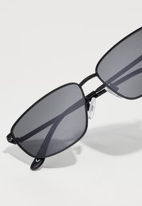 Schwarze Sonnenbrille mit übergroßen, kantigen Gläsern. Dünne, matte Rahmen mit Nasenpolstern und detailliertem Scharnier. Dunkel getönte Gläser.