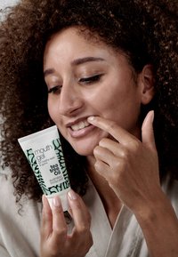 Mundgelrör med vit bas och grön botanisk design, märkt "tea tree oil," hålls av en person som undersöker sina tänder.