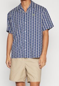 Košile s krátkým rukávem v námořnické modři s bílým geometrickým vzorem a malým zeleným logem na levé hrudi. Doplněné béžovými šortkami.