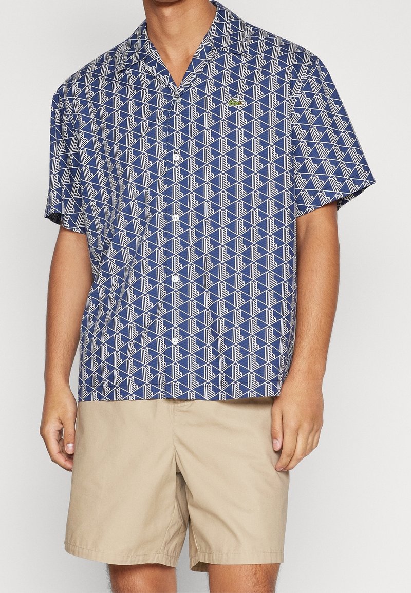Košile s krátkým rukávem v námořnické modři s bílým geometrickým vzorem a malým zeleným logem na levé hrudi. Doplněné béžovými šortkami.