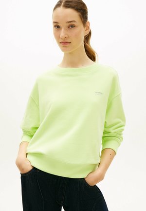 Giovane donna con capelli castano chiaro raccolti in una coda di cavallo, indossa una felpa verde chiaro e jeans scuri con le mani nelle tasche.