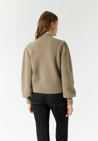 Een tan kleurige gebreide trui met pofmouwen, een geribbelde onderrand en kraag. Gecombineerd met donkere denim jeans die een relaxte pasvorm vanaf de achterkant laten zien.