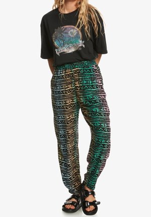 Kobieta w czarnym t-shircie z nadrukiem plażowej scenerii, kolorowych wzorzystych spodniach i czarnych sandałach z klamrą, stoi ze złożonymi za plecami rękami.