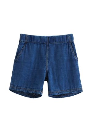 Młody chłopiec z kręconymi włosami noszący niebieską koszulę z krótkim rękawem na guziki, dopasowane szorty, granatowe skarpety z żaglówkami i wielokolorowe tenisówki.