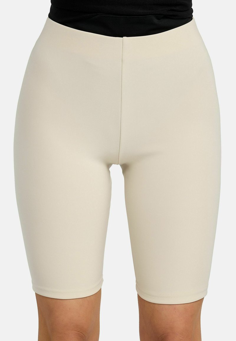 Shorts beige cintrés taille haute arrivant aux genoux, portés avec un haut noir sur une personne debout devant un fond uni.