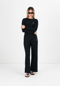 Fekete bordás, hosszú ujjú felső és széles lábú nadrág, kis logó részletel. A modell fekete napszemüveget és papucsot visel. Minimalista dizájn.