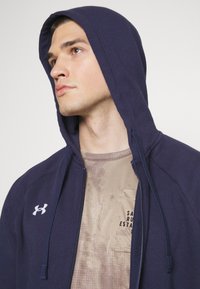 Donkerblauwe hoodie met rits in zachte stof, met een capuchon met trekkoorden en een wit logo. Draagover een lichtbruin gestructureerd shirt met een zak.